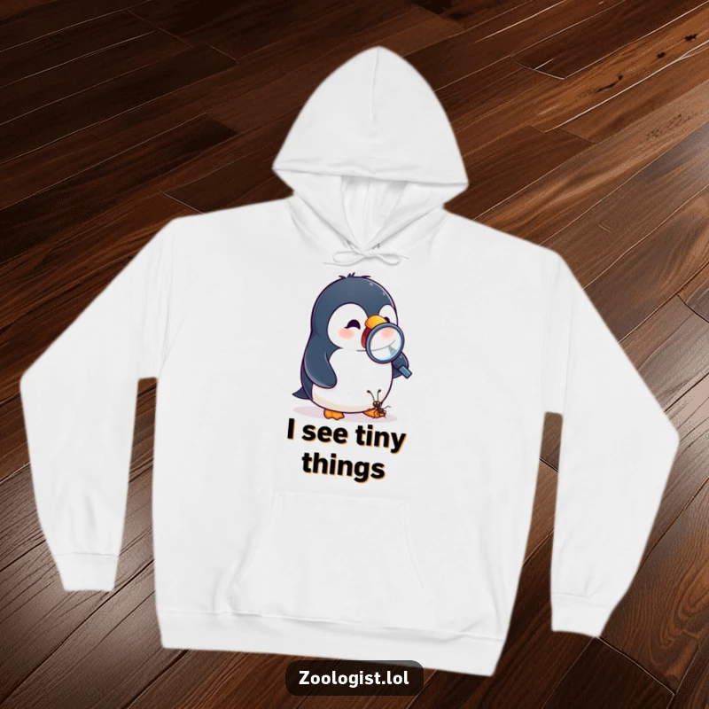 Funny hoodie depicting an excited penguin examining a tiny ant with a magnifying glass, great for cozy adventures.
