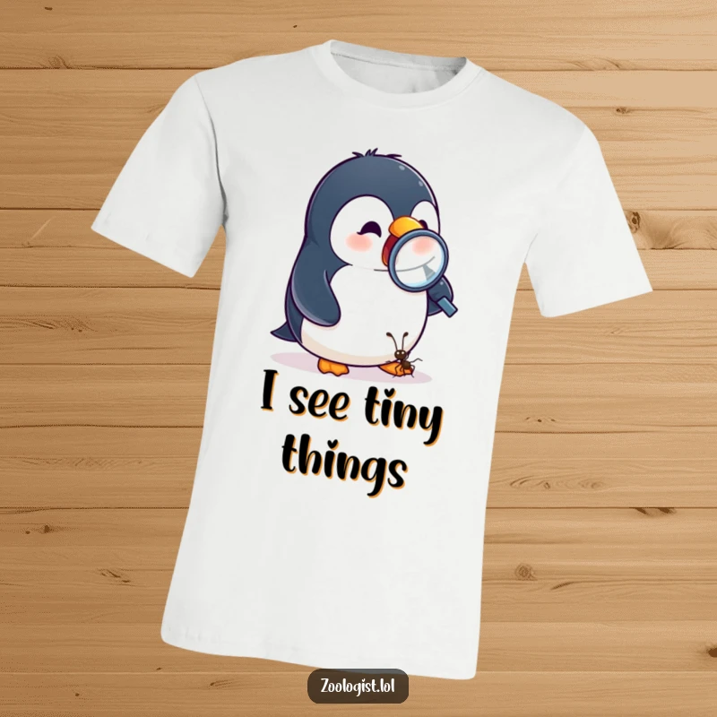 Funny t-shirt featuring an excited penguin closely observing a tiny ant with a magnifying glass, celebrating curiosity.